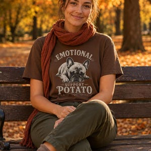 Może przedstawiać: Brązowa koszulka z napisem "EMOTIONAL SUPPORT POTATO" i grafiką buldoga francuskiego. Osoba ma na sobie szalik w kolorze rdzy i oliwkowe spodnie. Tło to park z jesiennymi liśćmi.