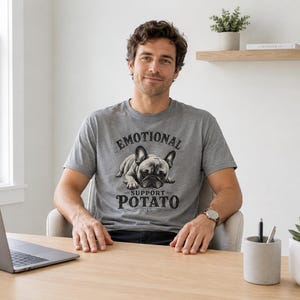 Może przedstawiać: Szara koszulka z napisem "EMOTIONAL SUPPORT POTATO" i grafiką buldoga francuskiego. Osoba siedzi przy drewnianym biurku z laptopem, pojemnikiem na długopisy i sukulentem w białej doniczce.