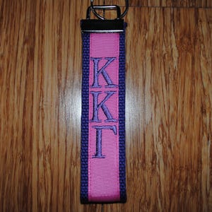 May include: A pink and purple key fob with the Greek letters KKT embroidered in purple. The key fob has a silver-colored metal ring at the top for attaching keys. The background is a wooden surface.