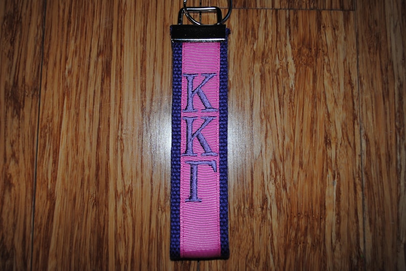 May include: A pink and purple key fob with the Greek letters KKT embroidered in purple. The key fob has a silver-colored metal ring at the top for attaching keys. The background is a wooden surface.