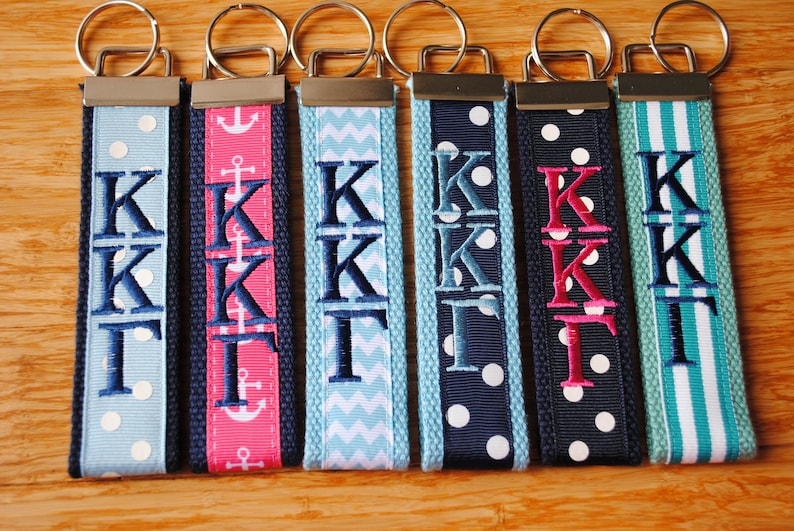 May include: A collection of personalized keychains with the embroidered letters "KKT". Each keychain features a different patterned ribbon in various colors, including blue, pink, and white, with a silver-colored metal clasp.
