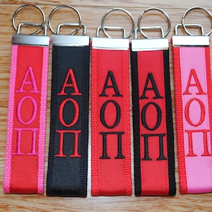 May include: Five keychains with red, black, and pink fabric straps. Each strap has the letters "AOP" embroidered in white or pink.