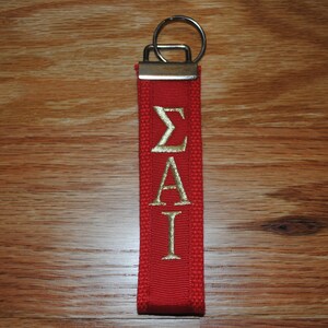 May include: Red fabric keychain with gold embroidered Greek letters &Sigma;&Alpha;&Iota;. The keychain has a silver metal ring and a rectangular metal piece at the top. The background is a wooden surface.