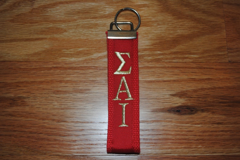 May include: Red fabric keychain with gold embroidered Greek letters &Sigma;&Alpha;&Iota;. The keychain has a silver metal ring and a rectangular metal piece at the top. The background is a wooden surface.