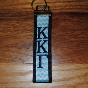 May include: A navy blue and light blue keychain wristlet with the Greek letters KKI embroidered in navy blue. The wristlet features a light blue and white chevron pattern and a silver key ring.