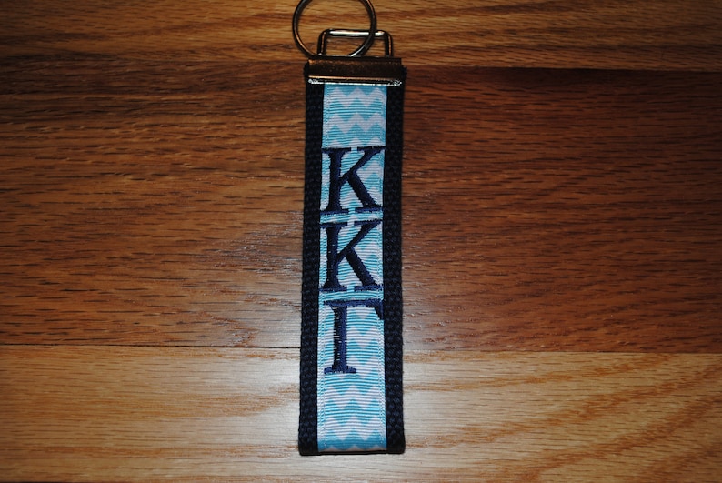 May include: A navy blue and light blue keychain wristlet with the Greek letters KKI embroidered in navy blue. The wristlet features a light blue and white chevron pattern and a silver key ring.