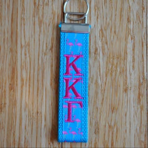 May include: A blue keychain with the Greek letters KKI in pink. The keychain has a silver metal ring and a pattern of pink flamingos. The background is a wooden surface.