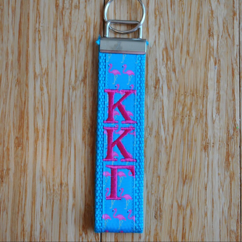 May include: A blue keychain with the Greek letters KKI in pink. The keychain has a silver metal ring and a pattern of pink flamingos. The background is a wooden surface.