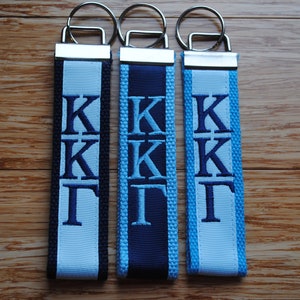 May include: Three keychains with embroidered Greek letters. Each keychain features a different color scheme: black and light blue, navy and light blue, and light blue. The keychains have silver-colored metal rings.
