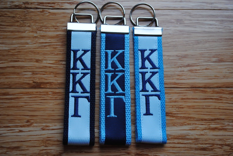 May include: Three keychains with embroidered Greek letters. Each keychain features a different color scheme: black and light blue, navy and light blue, and light blue. The keychains have silver-colored metal rings.