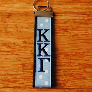May include: A blue and white polka dot keychain with the letters KKI embroidered in navy blue. The keychain has a silver metal clasp and a black border. The background is a wooden surface.