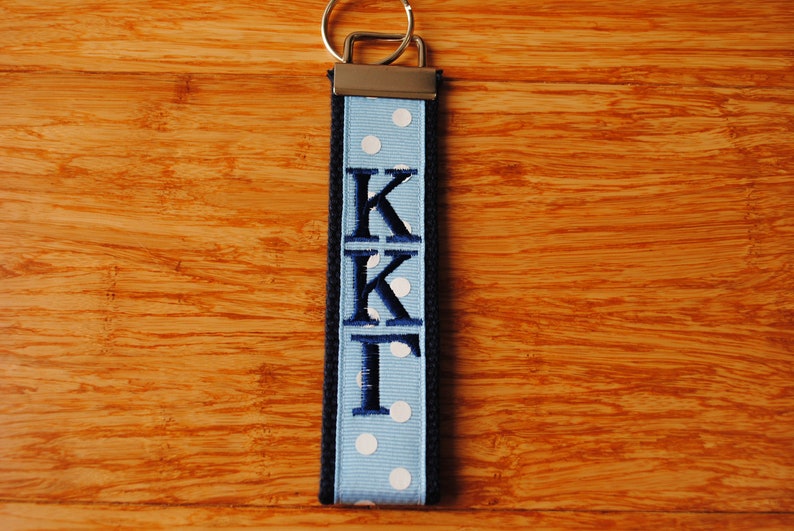 May include: A blue and white polka dot keychain with the letters KKI embroidered in navy blue. The keychain has a silver metal clasp and a black border. The background is a wooden surface.