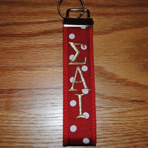 May include: Red keychain with white polka dots and gold embroidered Greek letters &Sigma;&Alpha;&Iota;. The keychain has a silver metal ring and a red fabric loop. The background is a wooden surface.