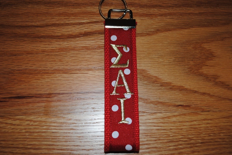 May include: Red keychain with white polka dots and gold embroidered Greek letters &Sigma;&Alpha;&Iota;. The keychain has a silver metal ring and a red fabric loop. The background is a wooden surface.