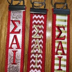 May include: Three red keychains with silver metal hardware. Each keychain features the Greek letters Sigma, Alpha, and Iota embroidered in red or gold. The keychains have decorative ribbon accents in silver, white, and gold.