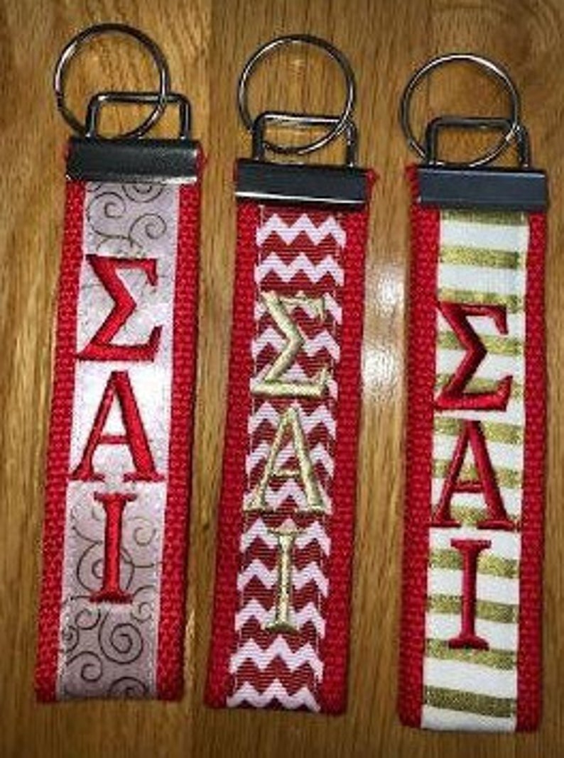 May include: Three red keychains with silver metal hardware. Each keychain features the Greek letters Sigma, Alpha, and Iota embroidered in red or gold. The keychains have decorative ribbon accents in silver, white, and gold.