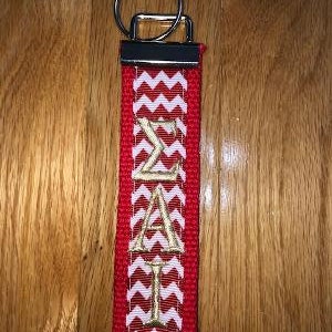 May include: Red and white chevron patterned keychain with gold embroidered Greek letters. The keychain has a silver metal ring and a red fabric loop. The letters read "&Sigma;&Alpha;&Iota;".