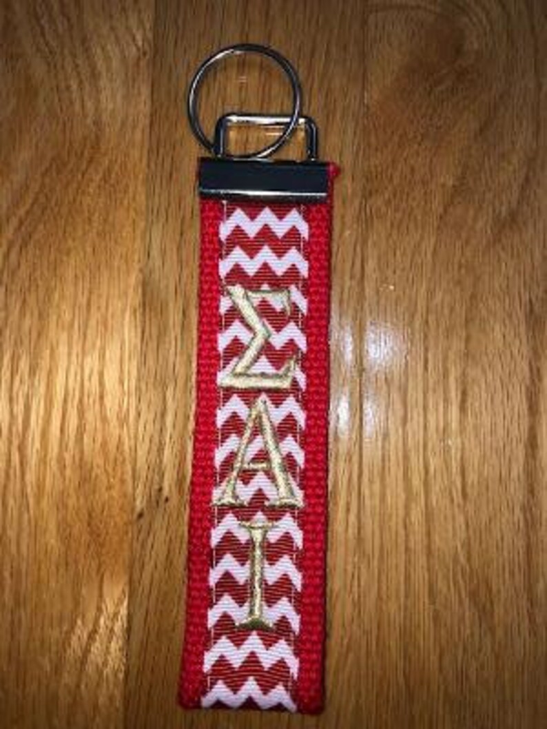 May include: Red and white chevron patterned keychain with gold embroidered Greek letters. The keychain has a silver metal ring and a red fabric loop. The letters read "&Sigma;&Alpha;&Iota;".