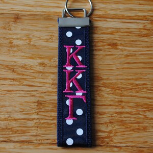 May include: Navy blue key fob with white polka dots and the embroidered letters "KKF" in pink. The key fob has a silver-colored metal ring.