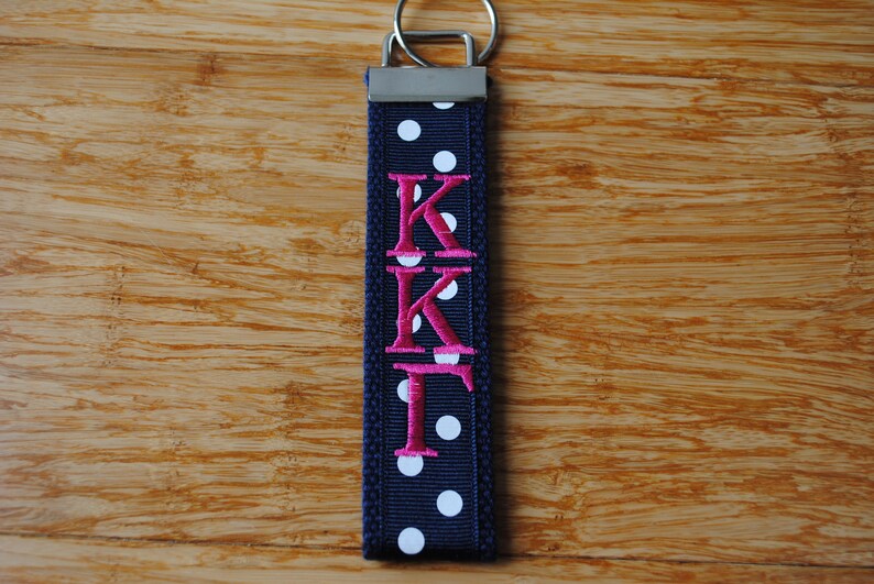 May include: Navy blue key fob with white polka dots and the embroidered letters "KKF" in pink. The key fob has a silver-colored metal ring.