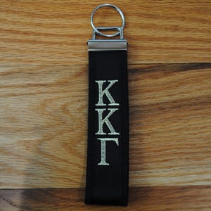 May include: A black fabric keychain with a silver metal ring and accents. The keychain features the embroidered letters "KK&Gamma;" in a silver color. The background is a wooden surface.