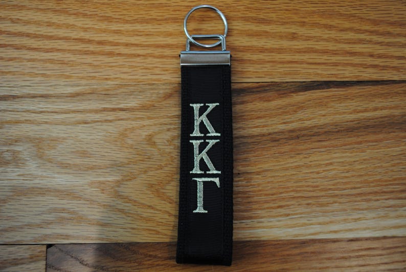 May include: A black fabric keychain with a silver metal ring and accents. The keychain features the embroidered letters "KK&Gamma;" in a silver color. The background is a wooden surface.