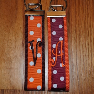 May include: Two orange and brown keychains with white polka dots. The keychains are personalized with the letters "V" and "A" in brown embroidery.