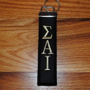 May include: Black keychain with gold embroidered Greek letters &Sigma;&Alpha;&Iota;. The keychain has a silver metal clasp and ring. The background is a wooden surface.