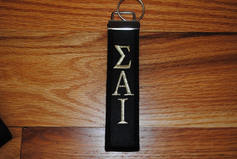 May include: Black keychain with gold embroidered Greek letters &Sigma;&Alpha;&Iota;. The keychain has a silver metal clasp and ring. The background is a wooden surface.