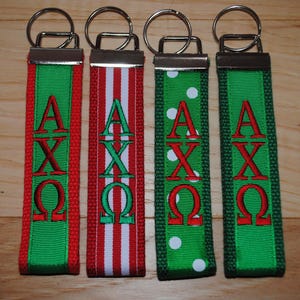 May include: Four keychains with metal rings. Each keychain has a green ribbon with red embroidered Greek letters. One keychain has red and white stripes, and another has white polka dots. The other two keychains are solid green.