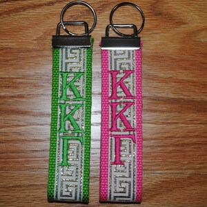 May include: Two keychains with a silver key ring and a fabric strap. One strap is green with white and gold Greek key design and green embroidered letters "KK&Gamma;". The other is pink with the same design and pink embroidered letters "KK&Gamma;".
