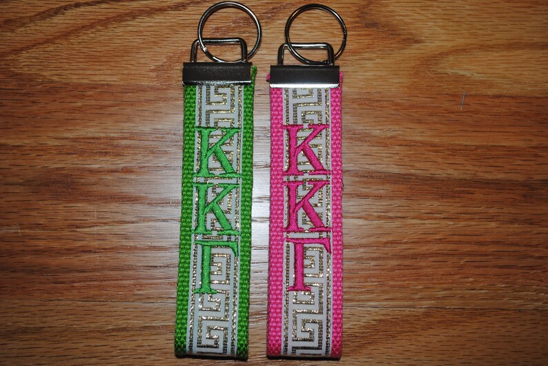 May include: Two keychains with a silver key ring and a fabric strap. One strap is green with white and gold Greek key design and green embroidered letters "KK&Gamma;". The other is pink with the same design and pink embroidered letters "KK&Gamma;".