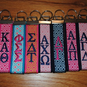 May include: A collection of eight keychains with fabric straps featuring embroidered Greek letters. The straps display a geometric pattern in various colors, including teal, pink, navy, and yellow. Each keychain has a silver metal ring.