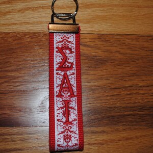 May include: A red and white fabric keychain with the Greek letters &Sigma;&Alpha;&Iota; embroidered in red. The keychain has a metal key ring and a decorative pattern along the edges.