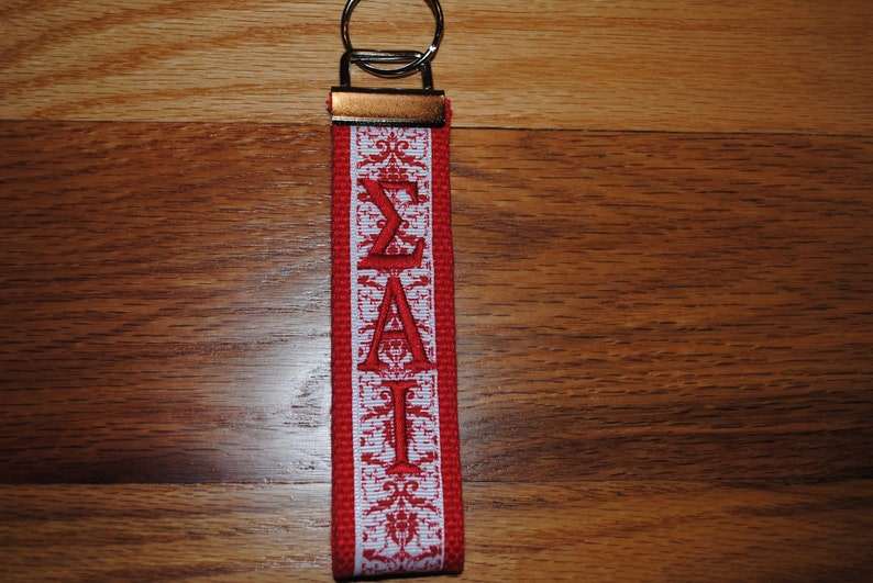 May include: A red and white fabric keychain with the Greek letters &Sigma;&Alpha;&Iota; embroidered in red. The keychain has a metal key ring and a decorative pattern along the edges.