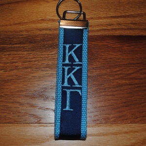May include: A blue and navy blue fabric keychain with the Greek letters Kappa Kappa Gamma embroidered in light blue. The keychain has a silver-colored metal ring and a dark wooden background.