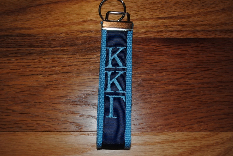 May include: A blue and navy blue fabric keychain with the Greek letters Kappa Kappa Gamma embroidered in light blue. The keychain has a silver-colored metal ring and a dark wooden background.