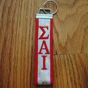 May include: A red and white keychain with the Greek letters &Sigma;&Alpha;&Iota; in red. The white section has gold stars. The keychain has a silver metal clasp and ring.