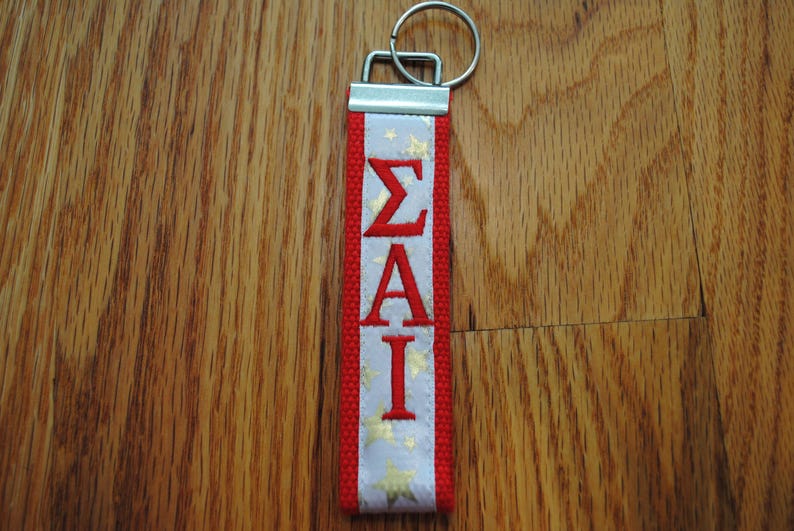 May include: A red and white keychain with the Greek letters &Sigma;&Alpha;&Iota; in red. The white section has gold stars. The keychain has a silver metal clasp and ring.