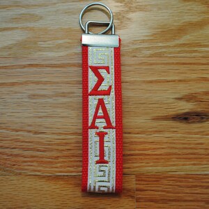 May include: A red and white keychain with the Greek letters &Sigma;&Alpha;&Iota; in red. The keychain has a silver metal ring and a red strap with white and gold accents. The background is a wooden surface.