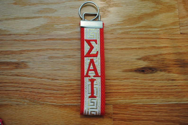 May include: A red and white keychain with the Greek letters &Sigma;&Alpha;&Iota; in red. The keychain has a silver metal ring and a red strap with white and gold accents. The background is a wooden surface.