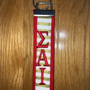 May include: A red, white, and gold key fob with the Greek letters &Sigma;&Alpha;&Iota; embroidered in red. The fob has a silver key ring and a black metal top. The background is a wooden surface.