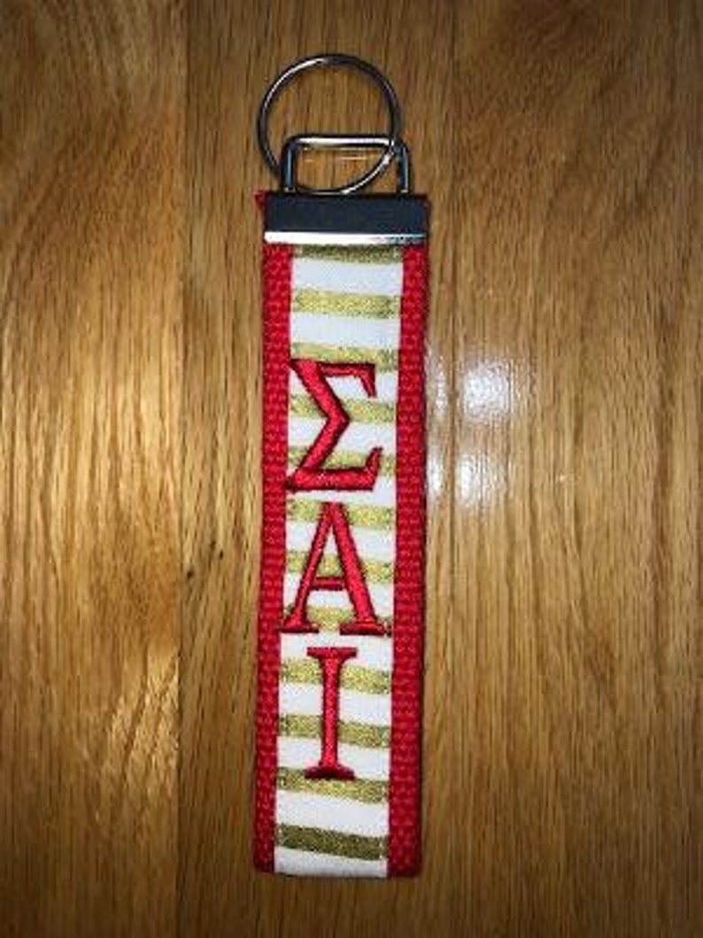 May include: A red, white, and gold key fob with the Greek letters &Sigma;&Alpha;&Iota; embroidered in red. The fob has a silver key ring and a black metal top. The background is a wooden surface.