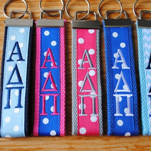 May include: Five fabric keychains with silver-tone metal hardware and split rings. Each keychain features embroidered Greek letters AΔΠ in various colors on ribbons with polka dot or chevron patterns in shades of blue, pink, and gray.