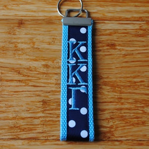 May include: A navy blue and light blue keychain with the embroidered letters "KKI" and white polka dots. The keychain has a silver metal ring for attaching keys.