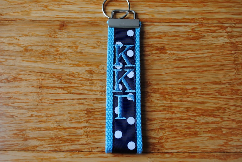 May include: A navy blue and light blue keychain with the embroidered letters "KKI" and white polka dots. The keychain has a silver metal ring for attaching keys.