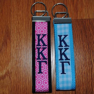 May include: Two keychains with metal rings. One has a pink and white geometric pattern with black trim, the other is light blue gingham. Both have the embroidered letters KKT in navy blue.