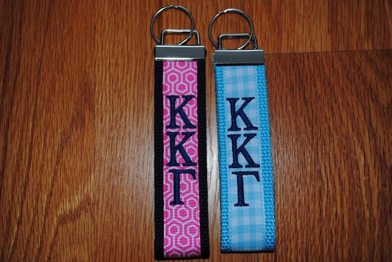 May include: Two keychains with metal rings. One has a pink and white geometric pattern with black trim, the other is light blue gingham. Both have the embroidered letters KKT in navy blue.