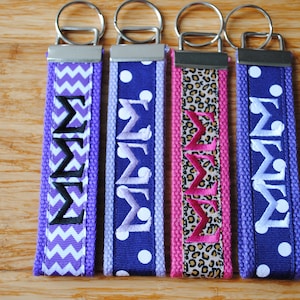 May include: Four purple and white patterned keychains with silver key rings. Each keychain has the Greek letters "ΣΣΣ" embroidered on it in different colors. The letters are embroidered in black, white, pink, and white.