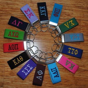May include: A collection of 14 colorful keychains with different Greek letter sorority and fraternity names embroidered on them. The keychains are arranged in a circle on a wooden surface.